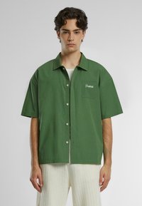 Camicia verde a maniche corte con colletto button-down, dotata di taschino sul petto e logo ricamato. Realizzata in un morbido tessuto di cotone.