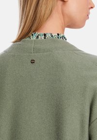 Grüner, gestrickter Pullover mit texturiertem Finish, Rüschekragen in Blau- und Schwarzmustern und einem Metall-Logo-Akzent auf der Rückseite.