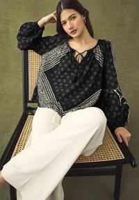 Blouse noire à motifs avec des manches volantes, décolleté à lacets, détails blancs contrastants. Le mannequin porte un pantalon blanc et est assis sur une chaise.
