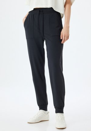 Zwarte trainingsbroek met elastische tailleband, trekkoord, twee voorkantenzakken en geribde manchetten, gecombineerd met witte sneakers tegen een lichte achtergrond.