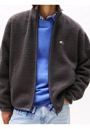 Mand iført en lynlåset koksgrå fleecejakke over en blå sweatshirt og ternet skjorte, kombineret med lyseblå revnede jeans.