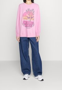 Rosa sweatshirt med grafisk design som visar träd och berg, i kombination med vida blå jeans och vita sneakers.