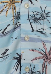 Tessuto di camicia azzurro chiaro con stampa tropicale che presenta palme, uccelli, montagne e una piccola barca a vela, con bottoni bianchi ben visibili.