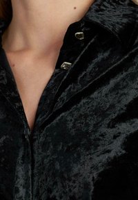 Camisa de terciopelo negro con cuello, textura sutil y herrajes decorativos plateados en los botones.