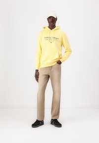 Gelber Hoodie mit Logo-Text, beigefarbene, gerade geschnittene Hose und schwarze Sneaker. Modell trägt eine weiße Kappe und steht vor einem schlichten Hintergrund.