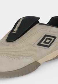 Μπεζ αθλητικό παπούτσι Umbro με υφή σουέτ, μαύρο λογότυπο και μαύρο προστατευτικό κάλυμμα που καλύπτει τα κορδόνια σε λευκό φόντο.