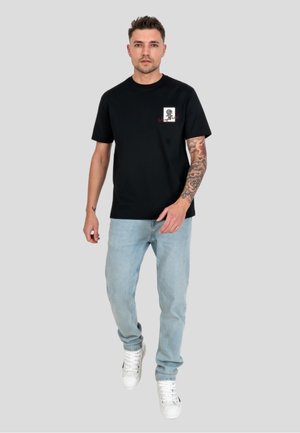 Hombre con tatuaje en el brazo derecho, vestido con camiseta negra con diseño gráfico, vaqueros azul claro y zapatillas blancas, caminando frente a un fondo gris liso.