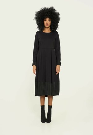 Zwarte lange mouw jurk met een geplooide rok en contrasterende glanzende zwarte zoom. Draag het met aansluitende zwarte enkellaarzen met een textuurstof.