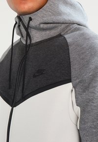 Gray and white zip-up hoodie featuring a high collar, drawstrings, and a black Nike logo on the chest. Made of blended fabric with smooth texture.