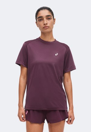Junge Frau mit zurückgebundenem dunklem Haar, die ein lila kurzärmeliges Sportshirt und passende Shorts trägt, steht vor einem schlichten Hintergrund.