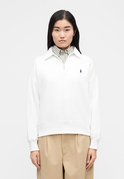 Jersey de algodón blanco con cuello de media cremallera, con un pequeño logo azul en el lado izquierdo. Combinado con una camisa de rayas verdes y blancas debajo.