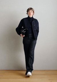 Nirhaļģa zila jūras pūķa jaka pāri melnai augstu aizsegšanai, kombinēta ar tumšām džinsu biksēm un baltām sporta kurpēm; rokās tur mazu melnu somiņu.