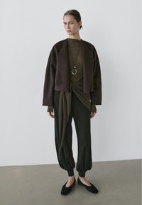 Brown cropped jacket, olive green top, dark loose-fitting trousers, and black flats. Features a twisted design and a circular necklace.