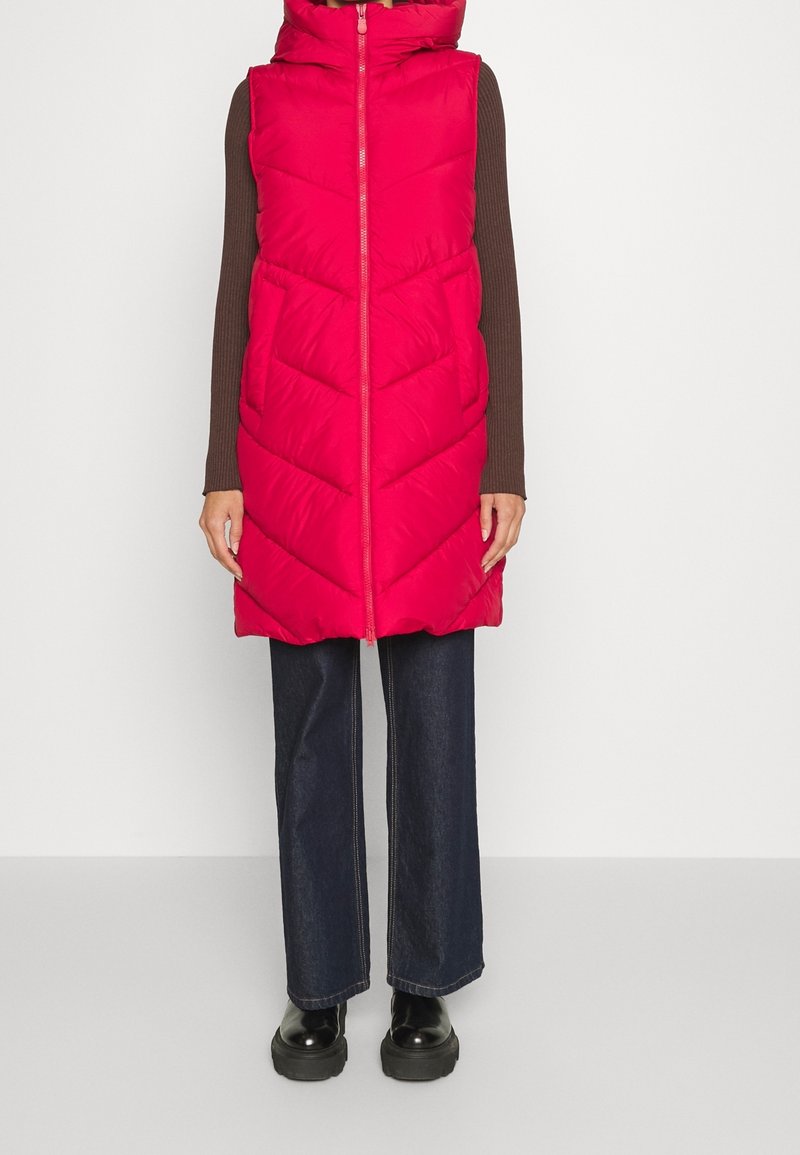 Roze puffer vest met een capuchon, voorzien van chevron-stiksel, ritssluiting en zijzakken, gedragen over een bruine longsleeve en donkere jeans.