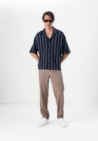 Camisa de botones de manga corta en color navy con rayas más claras, combinada con pantalones beige y zapatillas blancas, frente a un fondo liso.