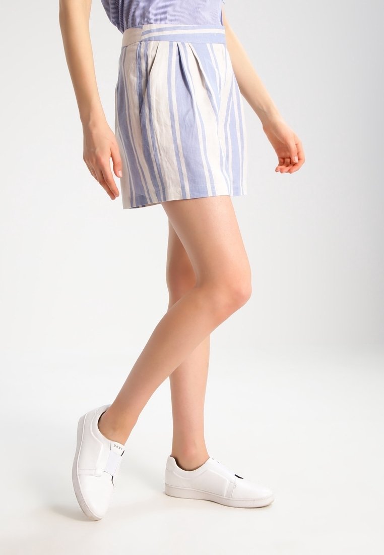 Femme portant un short rayé vertical bleu et blanc et des baskets blanches sans lacets sur un fond blanc uni.