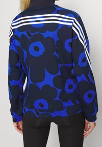Sudadera floral azul con fondo negro marino, que presenta tres franjas blancas en los hombros y un cuello alto. Hecha de una tela suave.