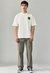 Hvid bomulds-t-shirt med korte ærmer og et sort firkantet logo, parret med olivengrønne cargo-bukser og sorte sneakers.