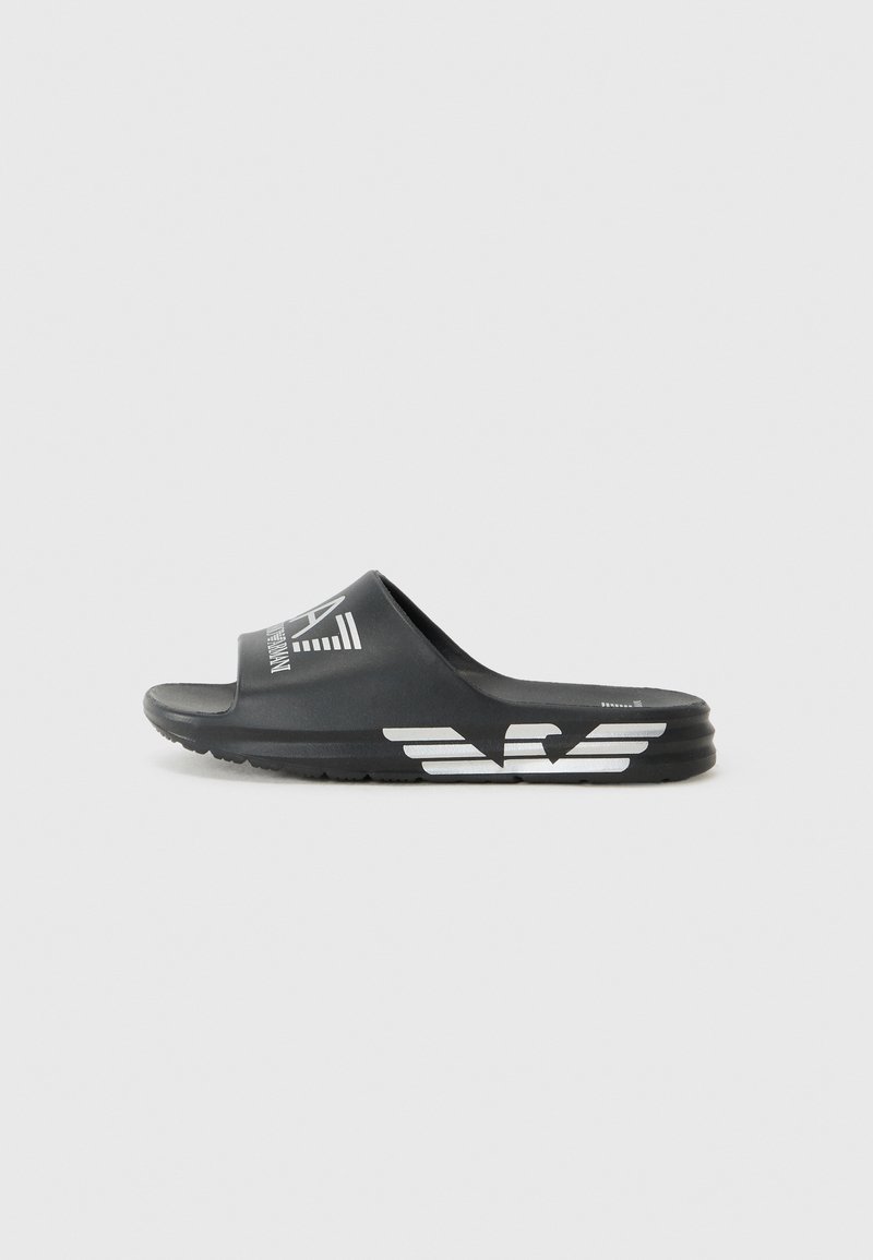 Black slip-on sandals made of textured rubber. Features a white logo design and a contoured footbed for comfort. Low-profile sole.