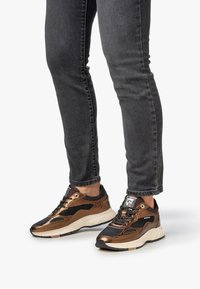 Jalat dalam jeans hitam ketat mengenakan sepatu sneakers perunggu dan hitam dengan sol putih, berdiri di latar belakang putih.