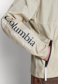 Ljusbeige Columbia-jacka med en texturerad yta. Vänster ärm har marinblå text "Columbia Sportswear Company." Dragkedjeficka vid midjan.