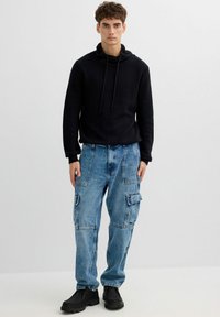 Sudadera negra texturizada con cuello ajustable con cordón, combinada con jeans cargo azul claro que presentan múltiples bolsillos y un ajuste relajado. Zapatos negros.