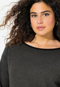 Pull gris foncé en matériau doux, avec un décolleté large et des accents côtelés. Cheveux bouclés visibles sur les côtés, texture de peau lisse.