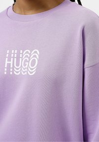 Sudadera lavanda con cuello redondo acanalado y corte holgado. Presenta un logo blanco "HUGO" en una fuente ondulada, fabricada con un tejido suave y texturizado.