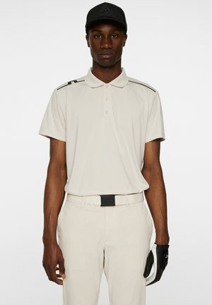 Vaaleanbeige polo-paita mustilla yksityiskohdilla, lyhyet hihat ja kaulus. Yhdistetty vaaleanbeigeihin housuihin ja mustaan lippikseen. Musta golf-käsine vasemmassa kädessä.