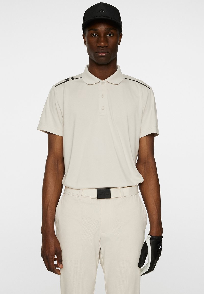 Ljusbeige polo skjorta med svarta detaljer, korta ärmar och krage. Matchad med ljusbeiga byxor och en svart keps. Svart golfhandske på vänster hand.