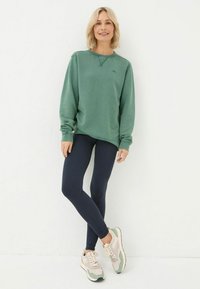 Green cotton sweatshirt with long sleeves, paired with dark leggings and beige sneakers featuring mint accents. Standing against a white background.