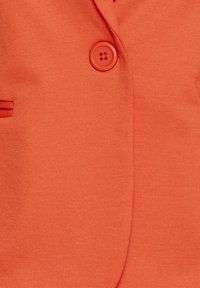 Üksik punane nupp kinnitatud ühevärvilise oranži bleiseri kangale, nähtava nööbiloa ja taskuõmblusega.