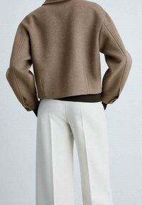 Veste courte marron avec un design structuré et un tissu texturé, assortie à un pantalon large crème, avec un accent noir à la taille.