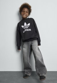 Sweatshirt noir avec logo Adidas blanc et trois bandes, associé à un jean gris à jambes larges et des chaussures noires en toile. Manches courtes et coupe décontractée.