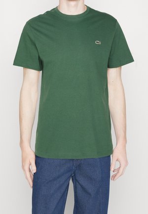 Homme portant un t-shirt vert uni à manches courtes avec un petit logo brodé sur la poitrine gauche et un jean bleu.