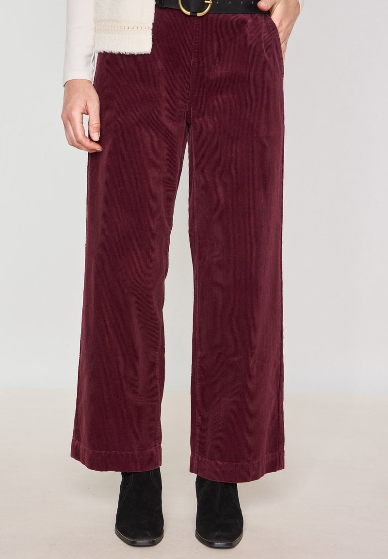 Pantaloni a gamba larga in corduroy bordeaux con una finitura testurizzata. Presentano un leggero risvolto all'orlo, abbinati a stivaletti neri per creare contrasto.