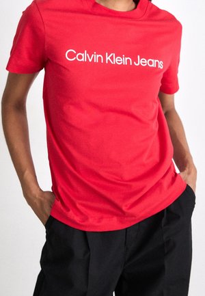 Osoba nosząca czerwoną koszulkę Calvin Klein Jeans z białym logo, w połączeniu z czarnymi spodniami, ręce w kieszeniach.