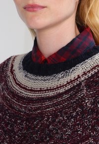Strickpullover in einem maroon- und cremefarbigen Farbschema, mit einem strukturierten Muster, Rundhals und einem geschichteten Karohemd in Rot und Marineblau.
