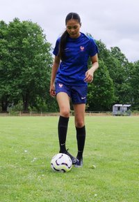 Blue soccer kit with short sleeves featuring a subtle pattern and pink accents, paired with black knee-high socks and black cleats, on grass.