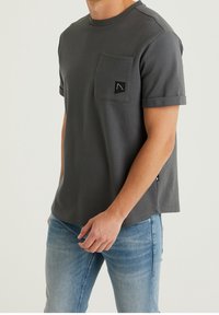 Uomo che indossa una camicia corta grigia scura con texture e tasca frontale, insieme a jeans azzurri chiari, in piedi contro uno sfondo chiaro e semplice.