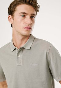 Polo shirt gris clair en tissu texturé, avec un col classique, une patte de boutonnage à deux boutons et un logo brodé sur la poitrine.
