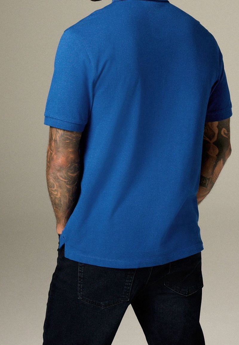 Homme aux bras tatoués portant un polo bleu uni à manches courtes et un jean foncé, de dos devant un fond neutre.