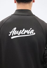 Οπίσθια όψη ενός ατόμου που φοράει μαύρο μπουφάν με τη λευκή γραφή «Austria» σε καλλιγραφικά γράμματα στο πάνω μέρος της πλάτης.