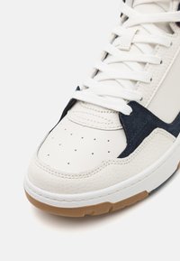 Sneaker, amely fehér bőrből készült felsőrésszel rendelkezik, kék velúr díszítésekkel, perforált orr résszel és gumi talppal. A fűzők fehérek.