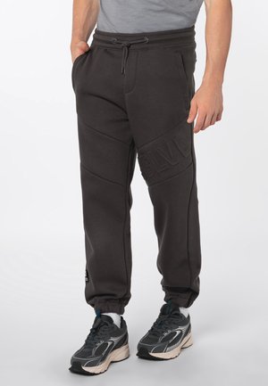 Mann trägt schwarze Jogginghosen mit Kordelzugbund, Seitentaschen, geprägten Buchstaben am Oberschenkel und schwarzen Sportschuhen mit weißen Socken.