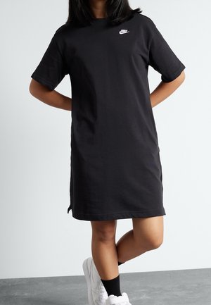 Person trägt ein schwarzes Nike T-Shirt-Kleid, schwarze Socken und weiße Sneakers, steht mit einem Bein über das andere geschlagen vor einem schlichten Hintergrund.