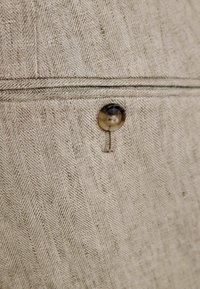 Tessuto beige con motivo a spina di pesce e un singolo bottone marrone cucito sopra una cucitura orizzontale.