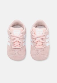 Un par de zapatos de bebé Adidas de gamuza rosa con franjas y cordones blancos, vistos desde arriba sobre un fondo blanco.