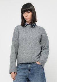 Donna che indossa un maglione grigio sopra una camicia a quadri blu con colletto, abbinato a jeans blu, in piedi con una mano in tasca su uno sfondo semplice.