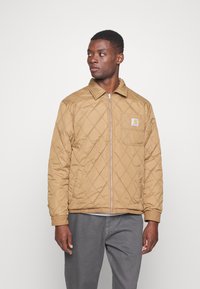Chaqueta acolchada marrón con cremallera frontal y un bolsillo en el pecho con un parche de logo, hecha de material ligero y con puños acanalados.